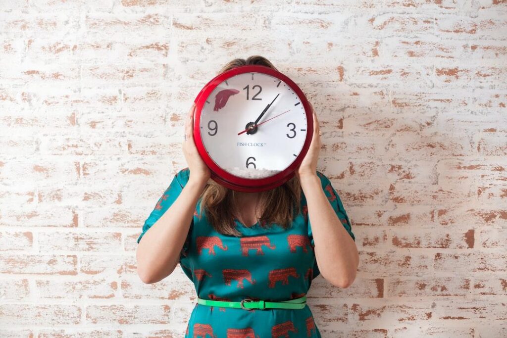 A person holds a big clock in front of their head, representing the manual tool that the best time clock for a small business should replace.